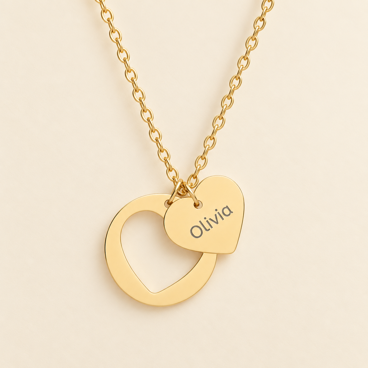 A gold necklace with a double-heart pendant, featuring one interlocking open heart and one small heart engraved with the name 'Olivia', shown on a light cream background.