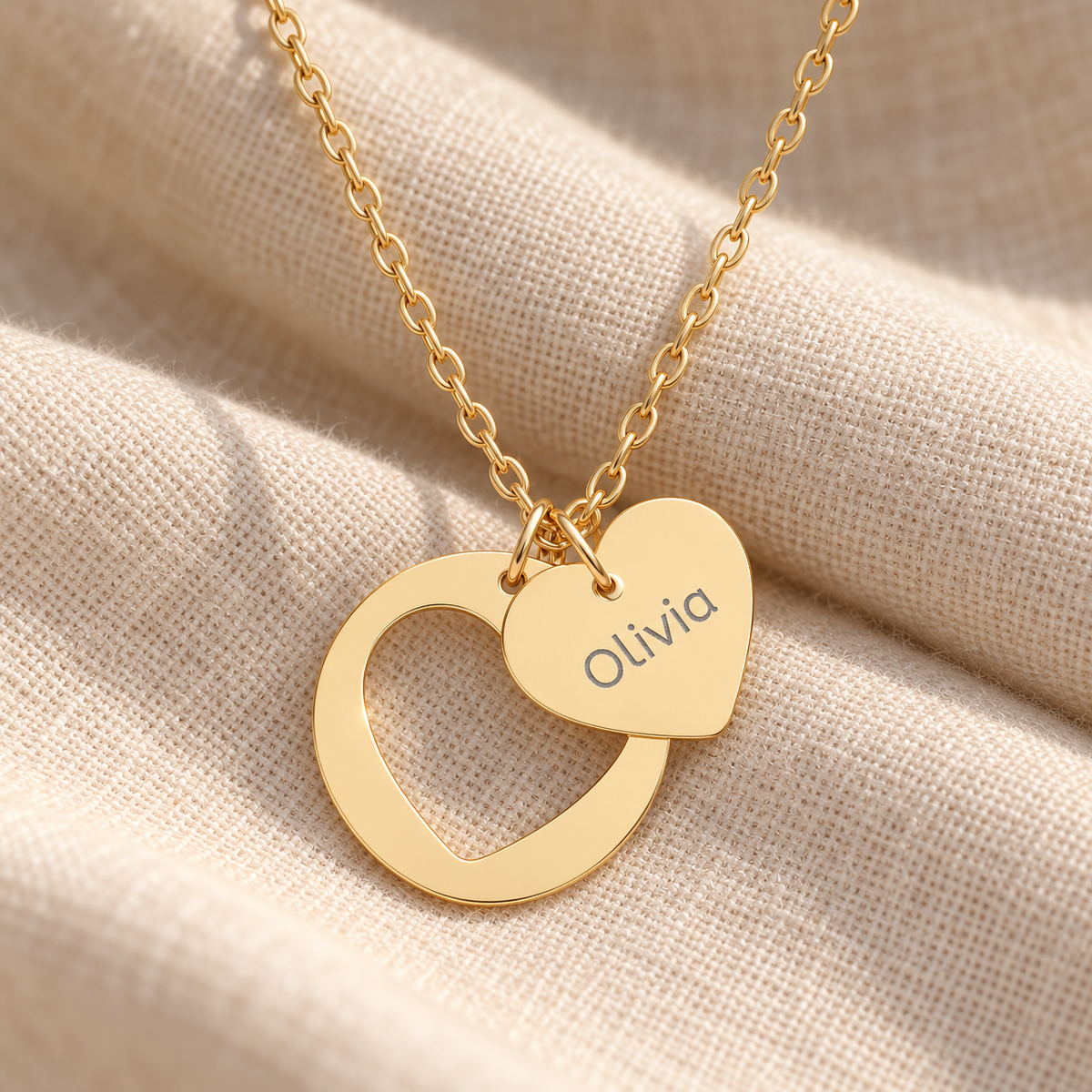 A gold necklace with two interlocking heart pendants, one engraved with the name 'Olivia', displayed on a textured beige fabric background.