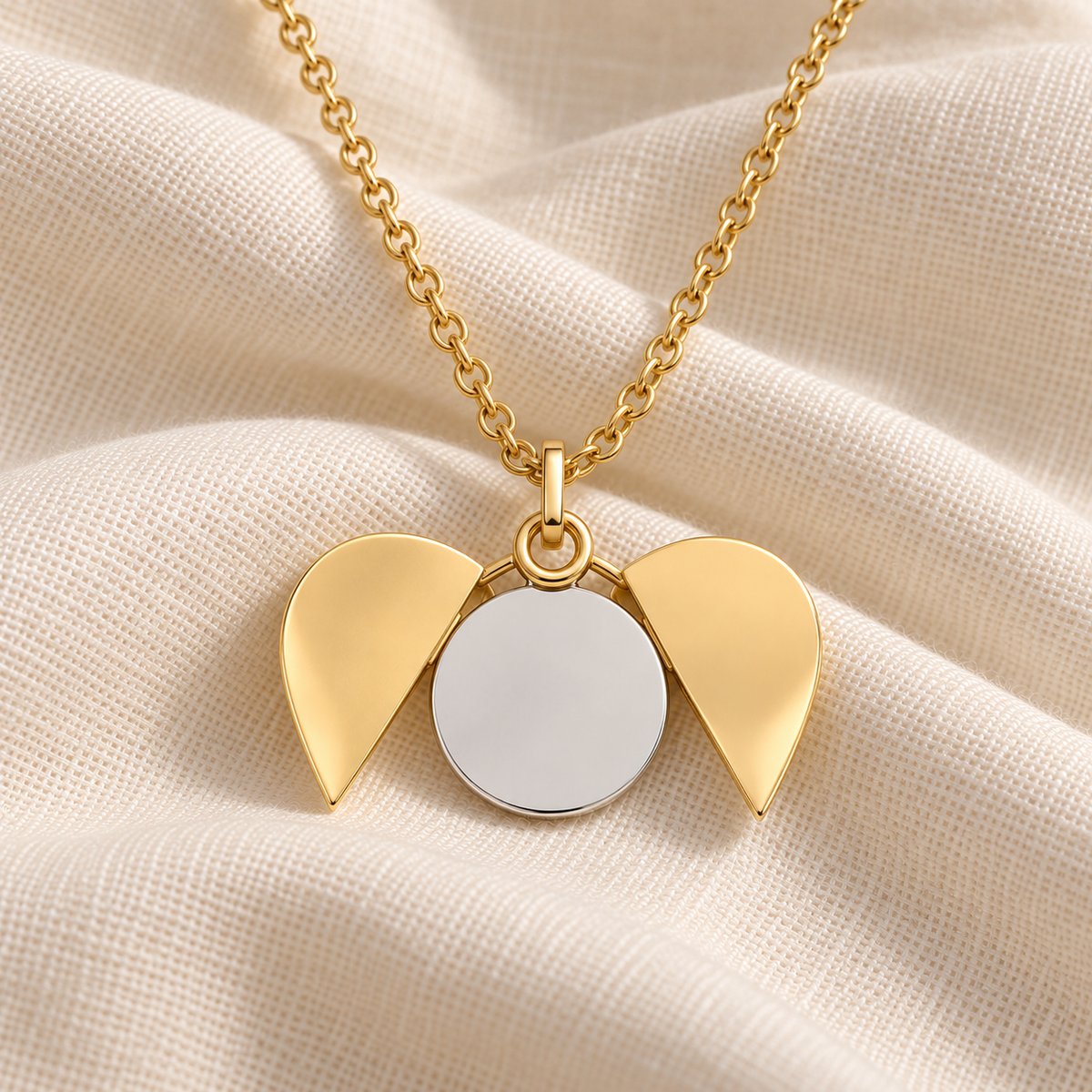 Gold angel wings pendant necklace with a blank silver circular center, displayed on a textured cream fabric background.