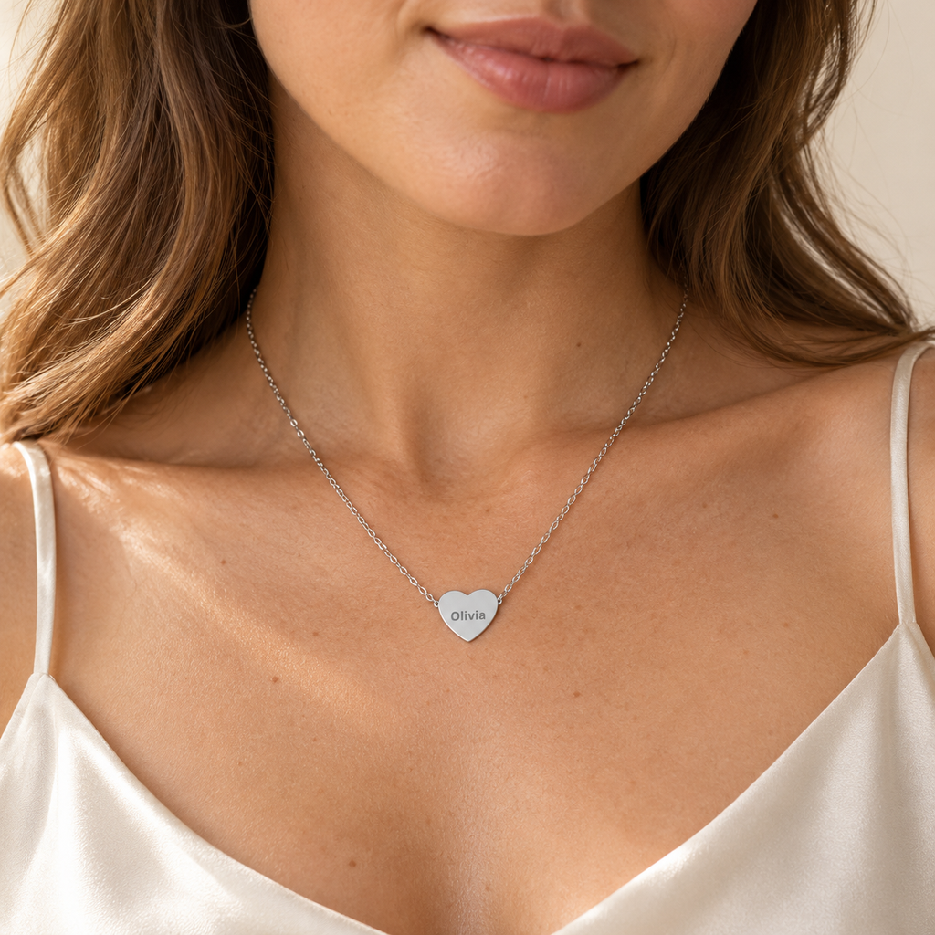 A close-up of a woman wearing a delicate silver chain necklace with a small heart-shaped pendant engraved with the name 'DISHA'. She is wearing a white satin camisole against a warm, neutral background.