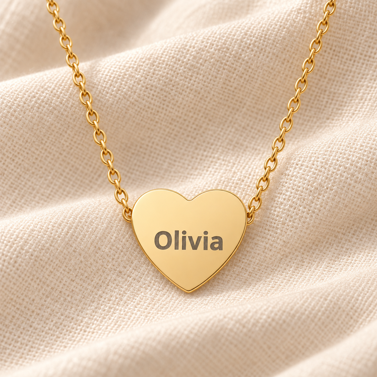 A gold heart-shaped pendant necklace engraved with the name 'Olivia' in dark lettering, resting on a textured beige fabric background.