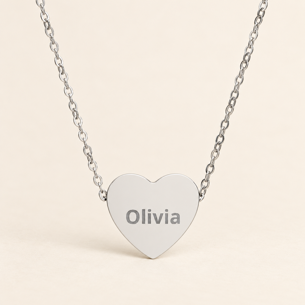 A silver heart-shaped pendant necklace engraved with the name 'Olivia', suspended from a delicate silver chain against a light beige background.