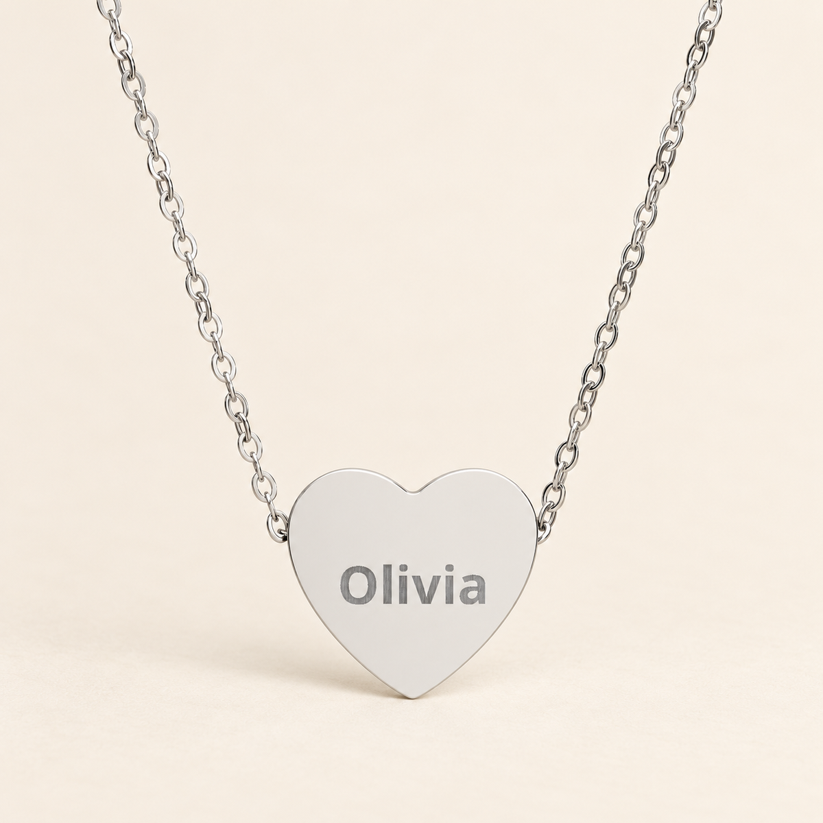 A silver heart-shaped pendant necklace engraved with the name 'Olivia', suspended from a delicate silver chain against a light beige background.