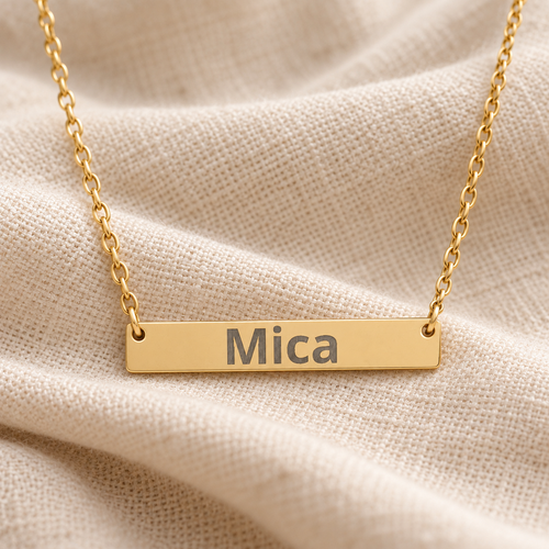Gold personalized bar necklace engraved with “Mica” resting on cream linen fabric.