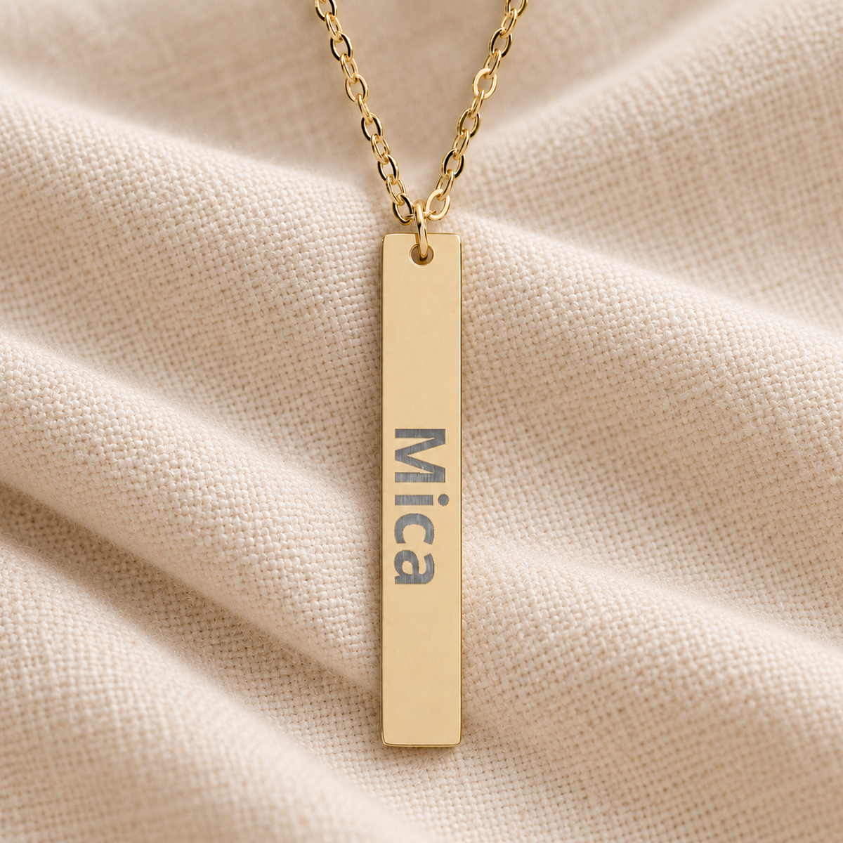 Gold-tone vertical bar necklace engraved with “Mica” resting on softly folded cream linen
